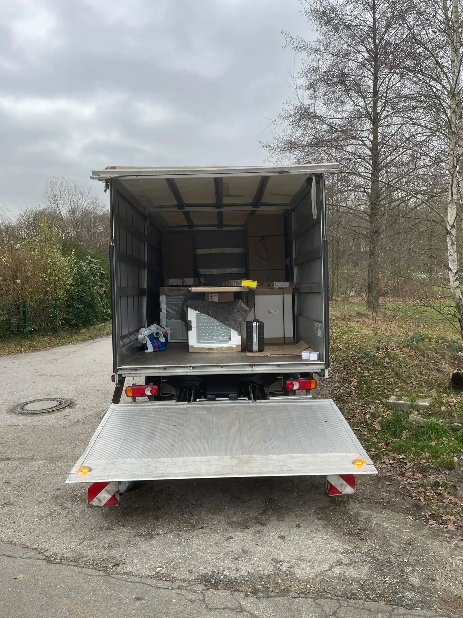 Transporter mit Kofferaufbau von hinten, Laderaum offen, Ladebordwand unten. Darin große Kartons, Geräte in Schutzverpackung unter einer Umzugsdecke, ein Werkzeugkoffer und eine Tasche.