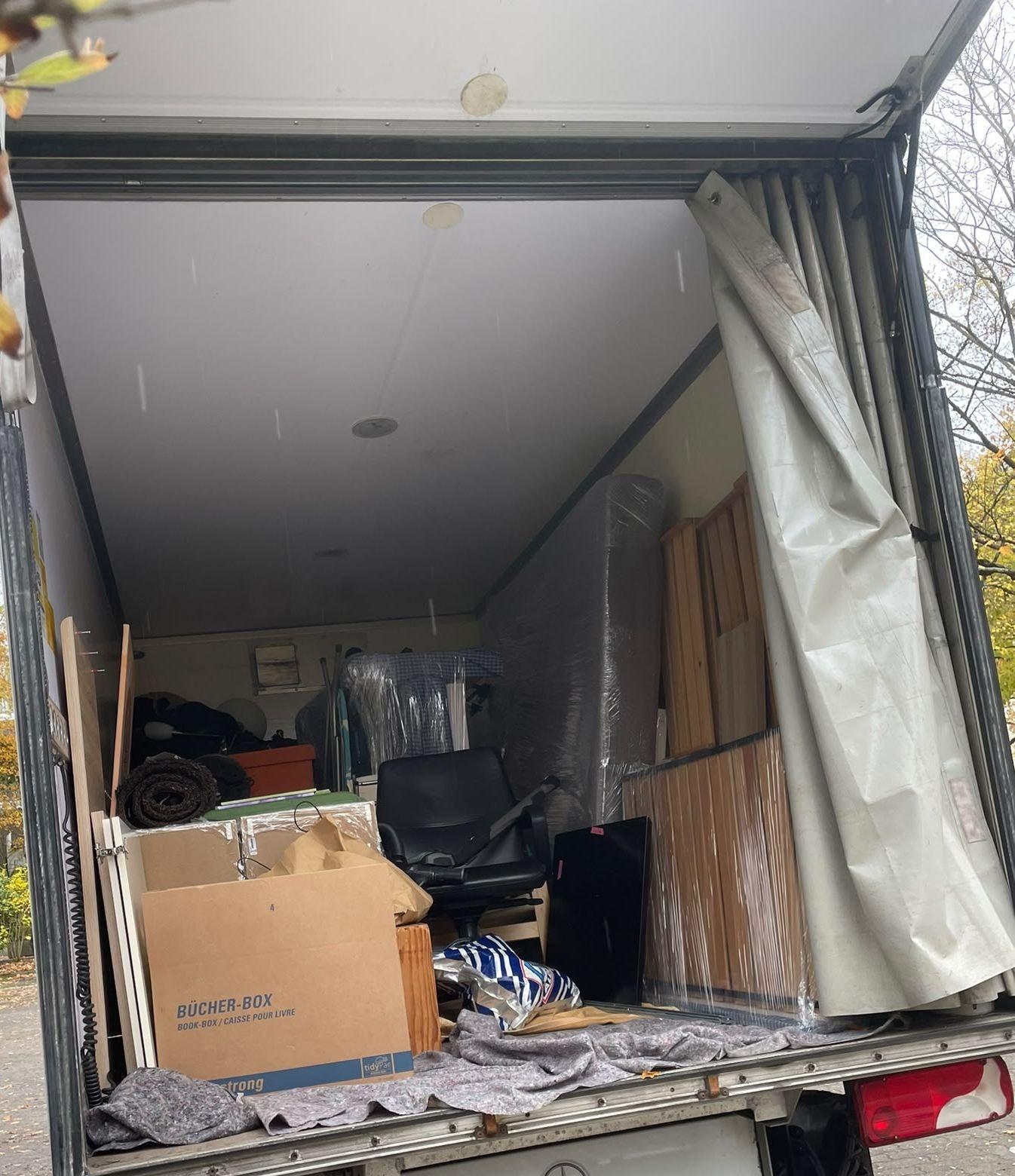 Geöffneter Laderaum eines Transporters mit Plane an der Seite: eine Matratze und Lattenroste in Folie, ein Bürostuhl, Kartons und Umzugsdecken auf dem Boden.